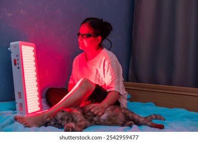 Senior woman treating sore leg with the red light therapy panel and wearing eye protection while petting a cat at home on the bed - Powered by Shutterstock