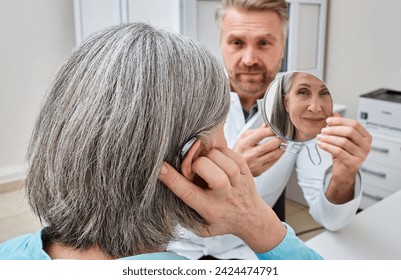 Senior woman trying on a hearing aid while looking into a mirror held by a hearing aid specialist in audiology clinic. Stylish hearing device - Powered by Shutterstock