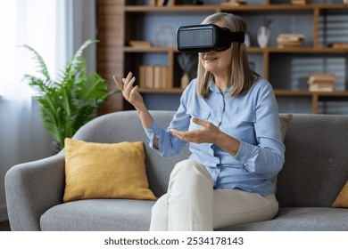 Senior woman uses VR headset seated on grey sofa, experiencing immersive virtual reality technology, expressing excitement, gesturing with hands, enjoying modern tech in home setting - Powered by Shutterstock