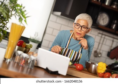 Senior woman using digital tablet at kitchen concentrate looking at tablet and search new recipes - Powered by Shutterstock