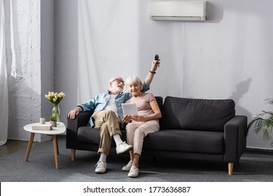 Senior woman using digital tablet near smiling husband switching air conditioner with remote controller