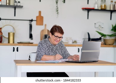 Senior Woman Using Laptop For Websurfing In Her Kitchen. The Concept Of Senior Employment, Social Security. Mature Lady Sitting At Work Typing A Notebook Computer In An Home Office.