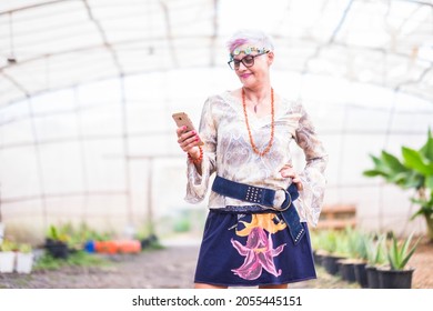 Senior Woman Using Mobile Phone While Posing With Hand On Hip Standing In Greenhouse. Elderly Woman Dreesed As Hippy Text Messaging On Mobile Phone. Caucasian Woman Enjoying At Greenhouse.