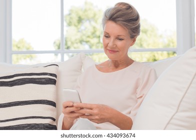 Senior Woman Using Mobile Phone While Sitting On Sofa. Older Woman Sitting On Sofa And Texting A Phone Message. Portrait Of A Beautiful Elderly Woman Learning To Use Smartphone.