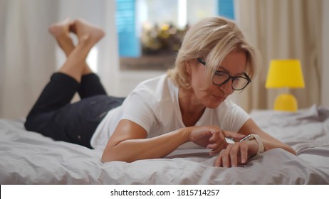 Senior woman using smartwatch relaxing on bed at home. Portrait of mature smiling lady using app or reading email on smartwatch lying on bed in cozy bedroom - Powered by Shutterstock