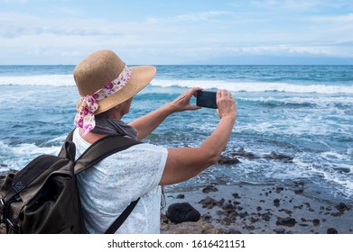 A Senior Woman In Vacation At The Sea Takes A Photo Of The Horizon Over Water. Casual Clothing With Backpack And Hat. Cloudy Sky