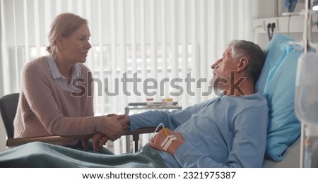 Senior woman visiting and cheering husband lying in bed at hospital ward. Sick aged male patient holding hands and talking to wife visitor in clinic room