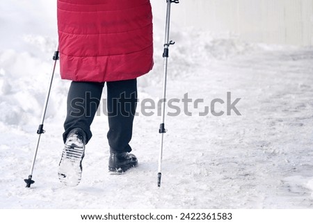Senior woman walking along slippery snowy sidewalk with walking poles. Old woman practicing Nordic pole walking, active lifestyle. Woman trekking with walking sticks in sunny winter day. 