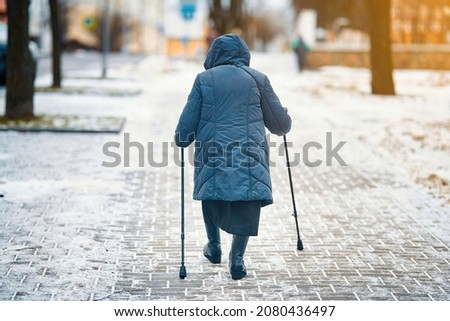 Senior woman with walking canes in hands walks along snowy walkway in winter city street. Old woman walking with cane, slippery sidewalk. Uncleaned pedestrian snowy road, risk of falls and injury