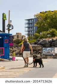 Senior Woman Walking With Her Black Dog On A City Street Of Victoria, BC, Canada-July 23, 2021. Travel Photo, Street View, Selective Focus, Concept Of The Photo Retierment.