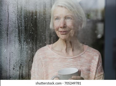 Senior Woman Is Watching The Rain From A Window In Her Home While Enjoying A Cup Of Tea.