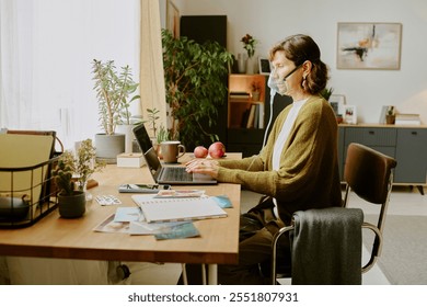 Senior woman wearing face mask sitting at desk working on laptop in home office, surrounded by plants and books, maintaining safety measures during pandemic - Powered by Shutterstock