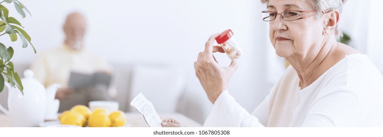 A Senior Woman Wearing Glasses Looking At A Transparent Bottle With Pills And Holding A Drug Information Leaflet. A Blurry Figure Of An Older Man Reading In The Background