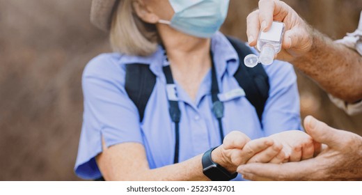 Senior woman wearing a mask receives hand sanitizer from a man. The woman, in a mask, enjoying outdoors. Senior woman using hand sanitizer. Senior couple enjoying outdoors adventure - Powered by Shutterstock