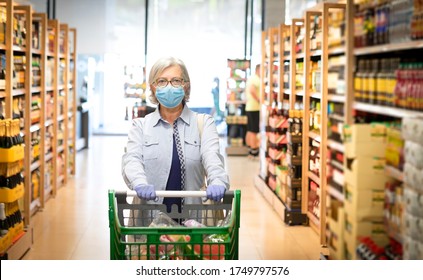 Senior Woman Wearing Mask And Rubber Gloves Pushes The Shopping Cart In A Supermarket, In The Wine Department - Active Elderly Pensioners, The Window In Background