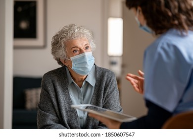 Senior Woman Wearing Safety Protective Mask At Home And Talking To Nurse. Back View Of Young Doctor Visiting Old Woman For Routine Health Checkup During Covid-19, Coronavirus And Flu Outbreak.