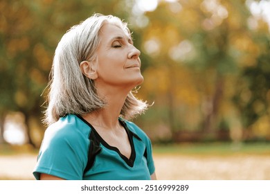 Senior woman wearing sportswear is enjoying a sunny autumn day in the park, closing her eyes and taking a deep breath of fresh air. Balance, harmony concept