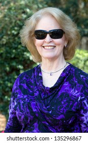 Senior Woman Wearing Sunglasses Stands Posing In The Backyard Outside Her Home And Smiles.