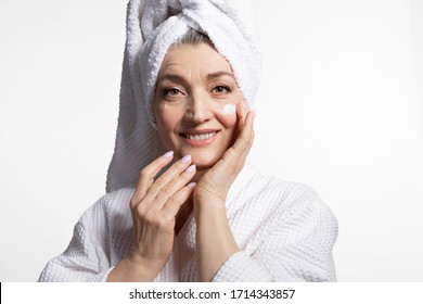 Senior woman wearing towel on head bathrobe applying moisturizing cream on face looking at camera. Mature woman after spa bath or shower on white. Health, skin care and cosmetology