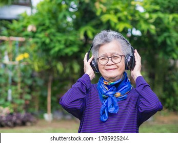 Senior Woman Wearing Wireless Headphone Listening To A Favorite Song While Standing In A Garden. Asian Old Woman, Short White Hair Are Enjoying The Music. Concept Of Old People And Healthcare