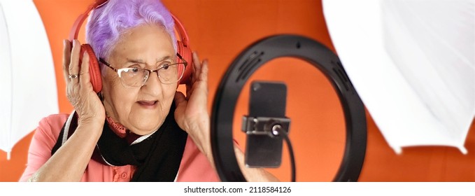 Senior Woman Web Influencer, With Purple Hair, Talks To Her Fans Via Smartphone With A Ring Of Light, On An Orange Background, With Her Hands On The Headphones