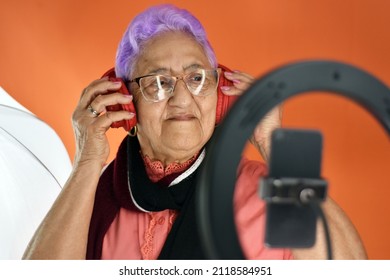 Senior Woman Web Influencer, With Purple Hair Listens Attentively To Her Fans By Smartphone In A Ring Of Light, On An Orange Background, With Her Hands On The Headphones