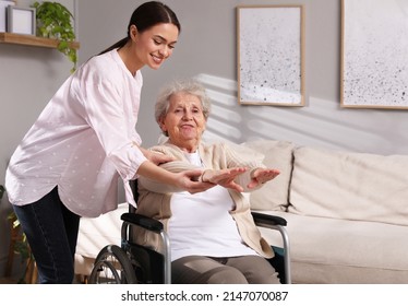 Senior Woman In Wheelchair Doing Physical Exercise And Young Caregiver Helping Her Indoors. Home Health Care Service