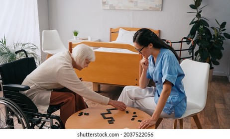 Senior Woman In Wheelchair Playing Dominoes With Young Nurse