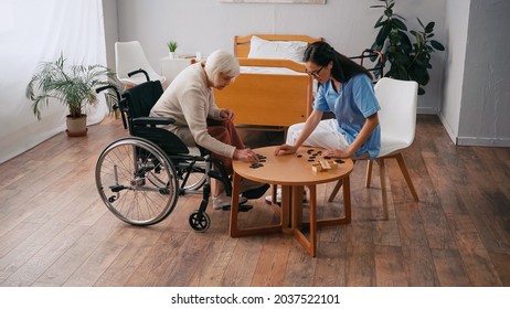 Senior Woman In Wheelchair Playing Dominoes With Brunette Nurse In Glasses