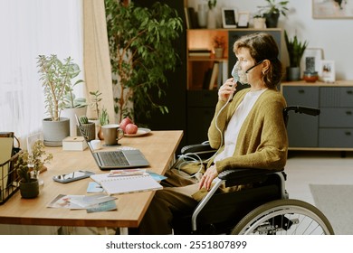 Senior woman in wheelchair using oxygen mask and working at desk with laptop and phone, surrounded by plants and office supplies - Powered by Shutterstock