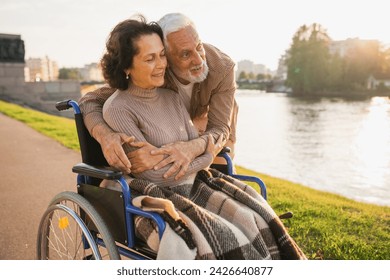 Senior woman in wheelchair walking with caregiver old man on road in park. Elderly family couple man supporting embracing woman in chair for people with disability outdoor. Rehabilitation - Powered by Shutterstock