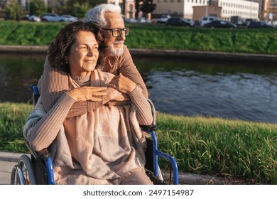 Senior woman in wheelchair walking with caregiver old man on road in park. Elderly family couple man supporting embracing woman in chair for people with disability outdoor. Rehabilitation - Powered by Shutterstock