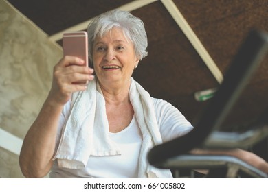 Senior Woman Working Out In Gym Sitting On Elliptical Machine And Typing On Smart Phone.