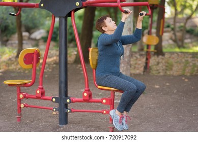 Senior Woman Working Out On The Sports Public Equipment In The Outdoor Gym.A Sportive Active Elderly Woman Doing Physical Exercise In A Park At Sunny Day.
