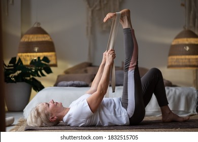 Senior Woman Working Out, Warming Up Using Yogic Belt, Lying In Yoga Supta Padangushthasana, One Leg Lift Exercise, Reclined Variation Of Extended Hand To Big Toe Pose At Home.