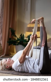 Senior Woman Working Out, Warming Up Using Yogic Belt, Lying In Yoga Supta Padangushthasana, One Leg Lift Exercise, Reclined Variation Of Extended Hand To Big Toe Pose At Home.