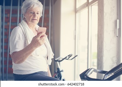 Senior Woman Workout In Gym. Woman Using Smart Phone On The Elliptical Machine.