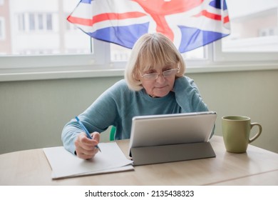 Senior Woman Writes New English Words During Distant Learning With Laptop, Language Classes For Ukrainian Refugee For Better Adaptation Abroad, British Flag On The Window, Social Distance Education