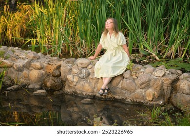 Senior woman in yellow dress sitting on a stone wall by a pond in a park. Outdoor nature portrait photography. Relaxation and peaceful lifestyle concept. A female person aged 50 years - Powered by Shutterstock