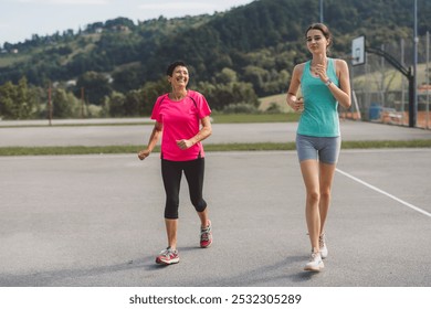 A senior woman and a young woman jogging together on an outdoor track, both smiling and enjoying their exercise. The background features green hills and a clear sky. - Powered by Shutterstock