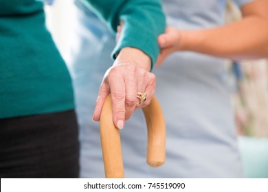 Senior Woman's Hands On Walking Stick With Care Worker In Background