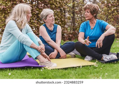 Senior Women Chatting After Yoga Session - Three senior women sit on their yoga mats outdoors on the grass, chatting together after a workout session. - Powered by Shutterstock