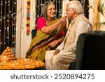 Senior women dressed feeding sweets to cheerful husband while sitting on chairs and celebrating Diwali festival together at home