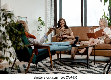 Senior Women Spending Time Together At Drawing Room. Blond Woman Holding Book. Red Haired Woman Stretched Legs On Coffee Table. Brunette Woman Drinking Red Wine. Natural Ageing. Diversity And Equity. 