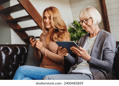 Senior women using digital tablet and middle aged woman, her daughter, mobile phone while relaxing on sofa at home. Social media, addiction to gadgets. Active seniors - Powered by Shutterstock