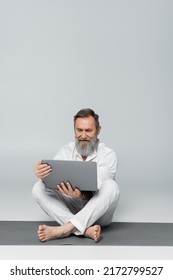 Senior Yoga Coach With Laptop Sitting On Yoga Mat In Easy Pose On Grey