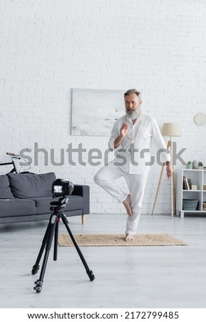senior yoga coach showing tree pose and prana mudra gesture in front of digital camera