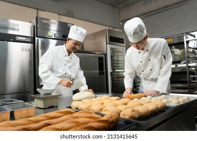 A senior and a young Asian male bakers in a white chef dresses and hats standing cutting, weighing, and kneading the dough on a counter with many baked and unbaked bread on trays at a bakery kitchen.
