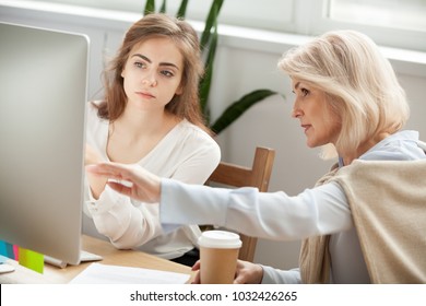 Senior and young female colleagues discuss online project look at pc screen, older mentor teaches young woman explains corporate software work, aged executive helps intern, teamwork on computer task