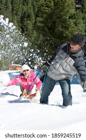 A Senior-adult Man Playfully Pulling His Cheerful Wife Through The Snowy Woods While She Is Sitting On A Sled And Throwing Snow In The Air Outside A Cabin